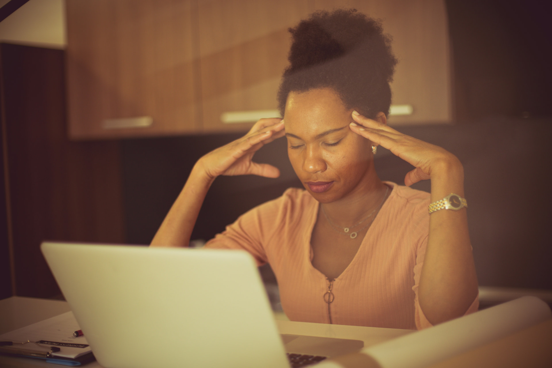An educator reflecting with hands at her temples while working on a laptop, illustrating the pressure of systemic workforce strain